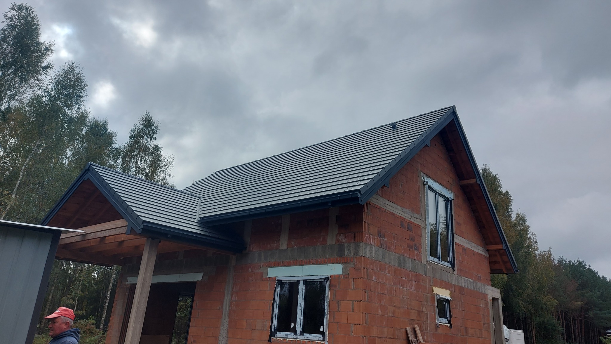 Surowy budynek z ciemnoszarą dachówką karpiówką i czarnymi rynnami, widok z dołu na tle pochmurnego nieba i drzew. Częściowo widoczna osoba w czerwonej czapce.