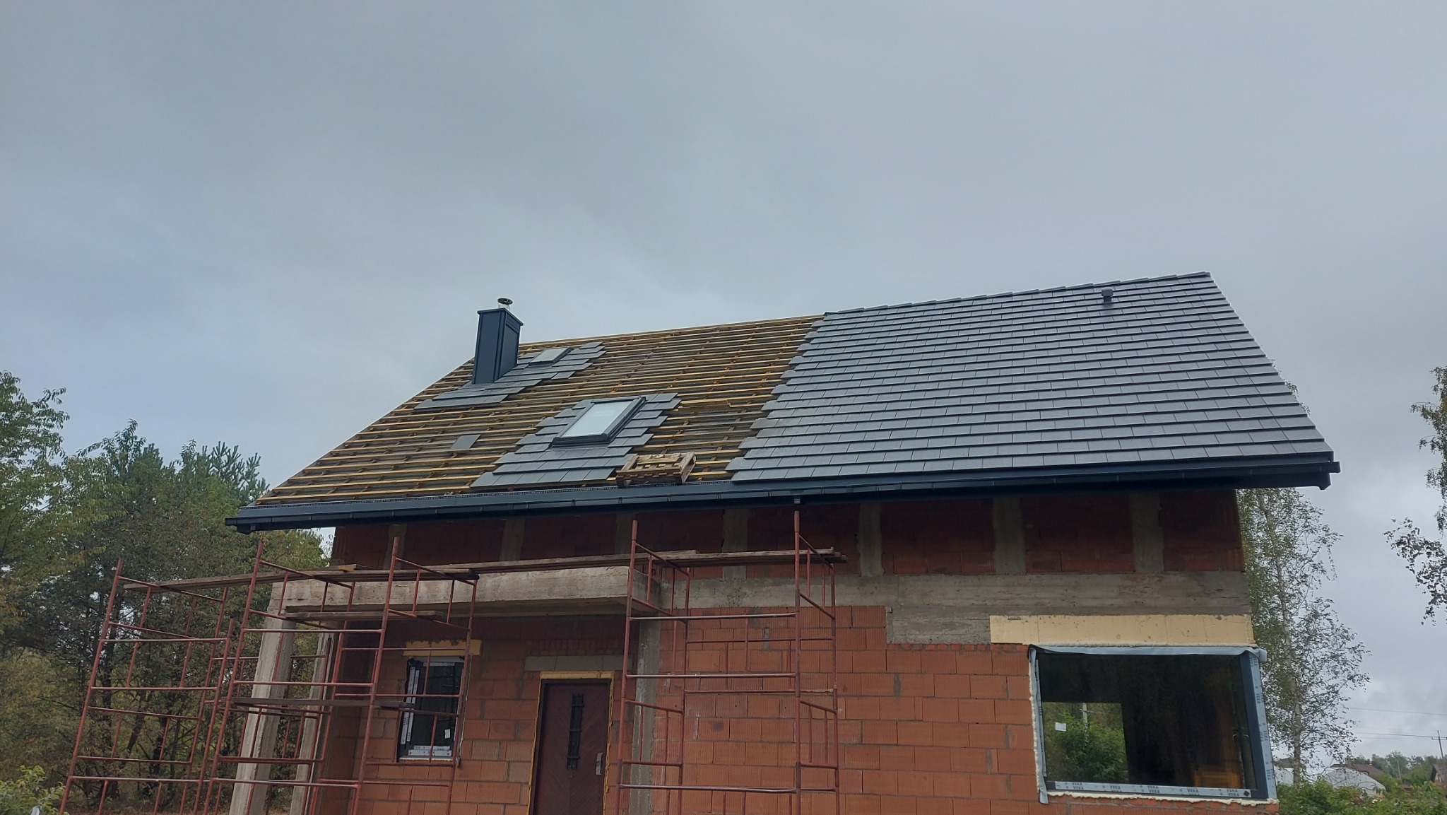 Dom w trakcie krycia dachu szarą dachówką. Widoczna więźba, komin i rusztowanie. Część dachu już pokryta, druga w trakcie prac. Okno dachowe zamontowane.