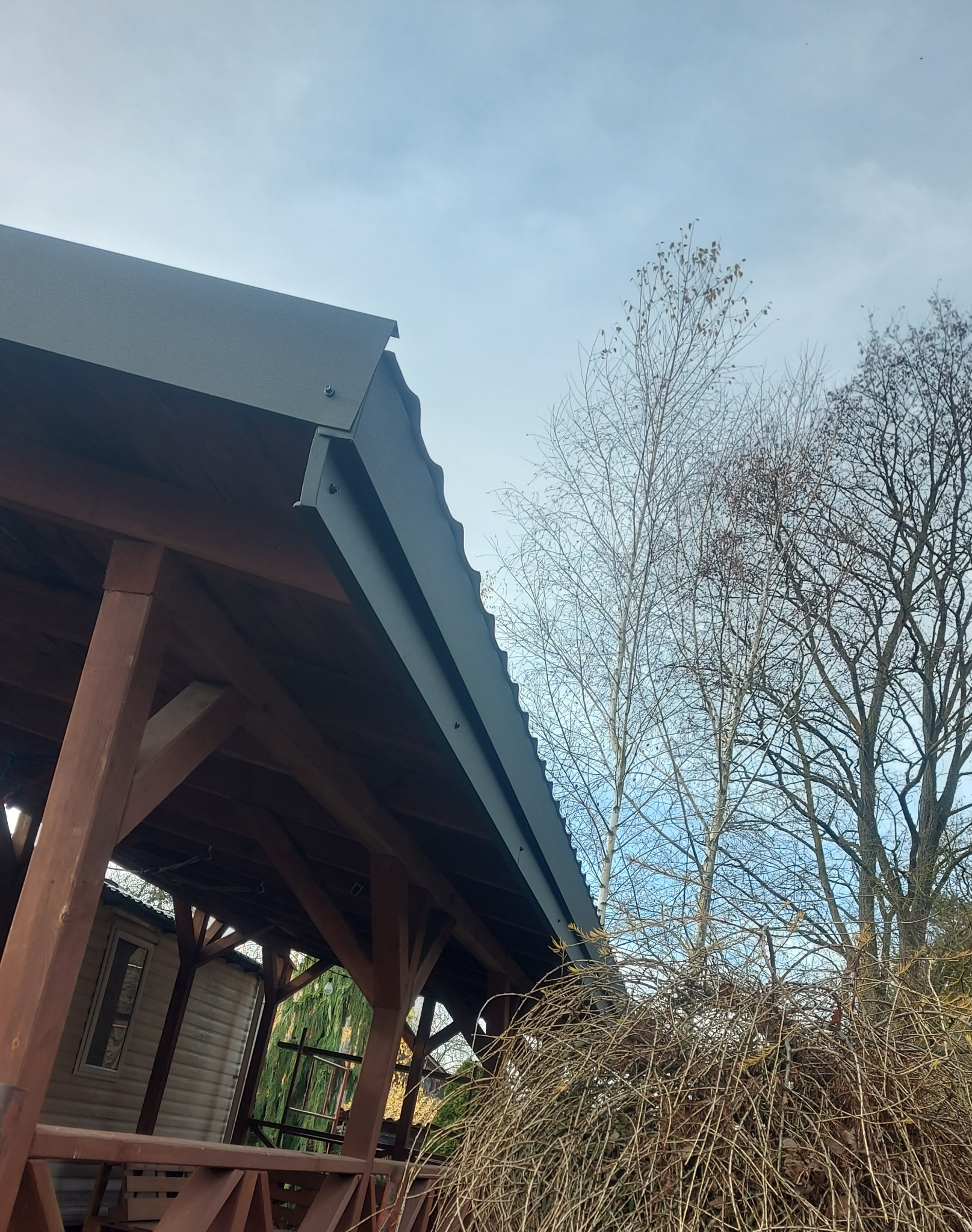 Zadaszenie altany z szarej blachy trapezowej, widoczne mocowania. W tle błękitne niebo i drzewa bez liści. Ujęcie z dołu, podkreślające konstrukcję dachu.