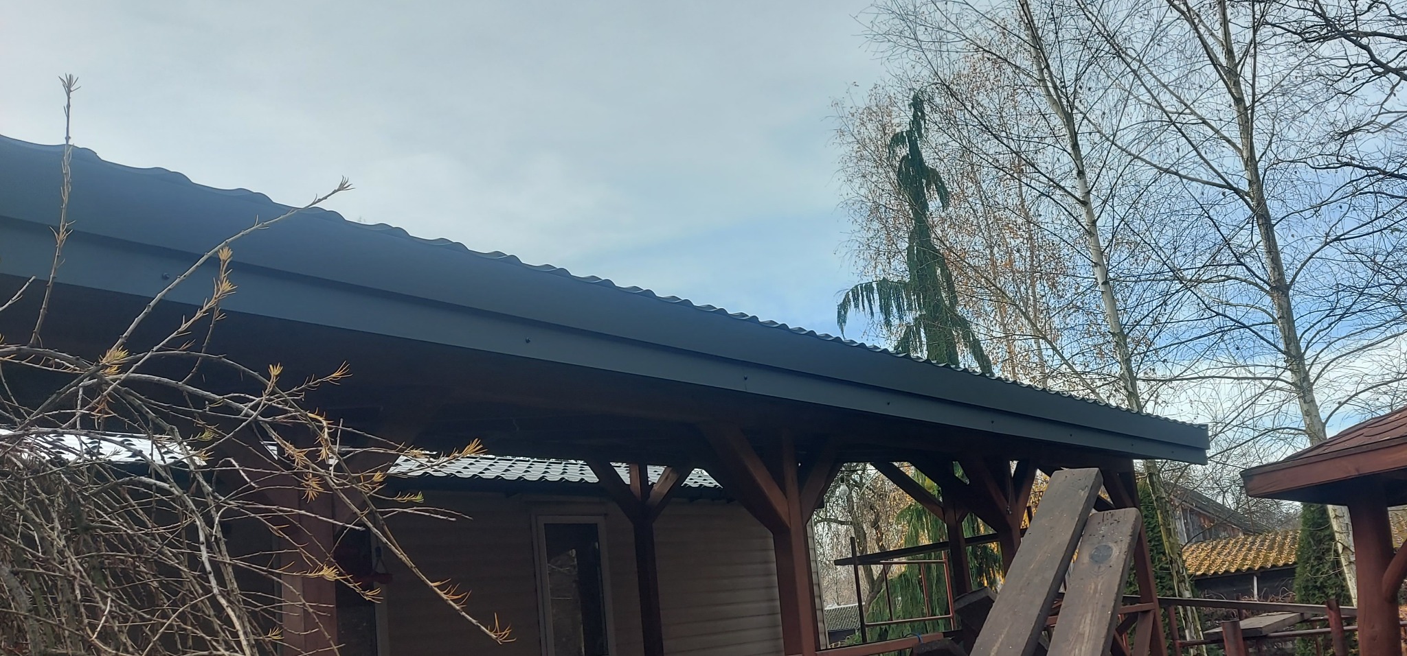 Zadaszenie tarasu z ciemnym, falistym pokryciem dachowym, drewnianą konstrukcją i widocznymi elementami montażowymi. W tle drzewa i błękitne niebo.