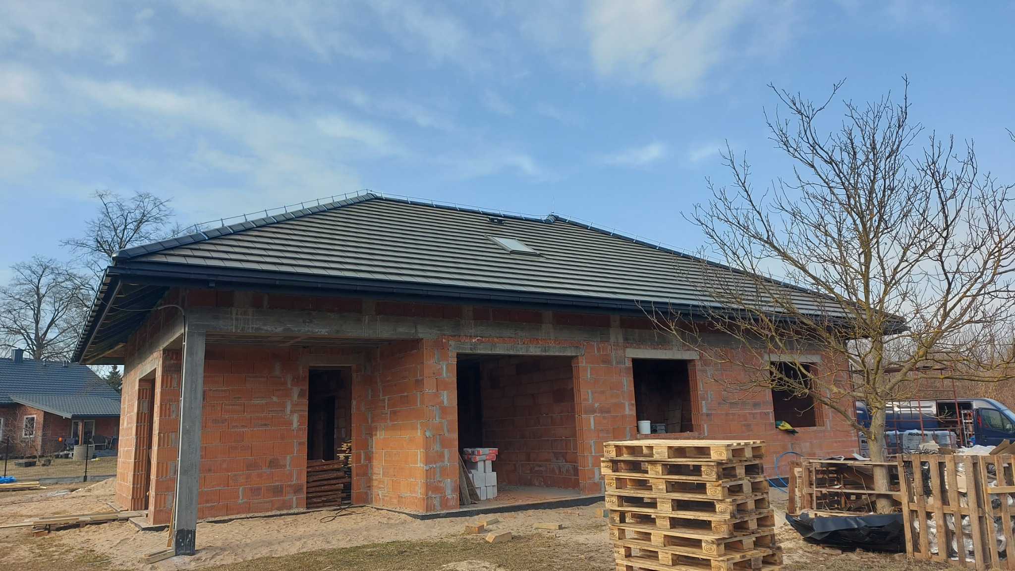 Dom w trakcie budowy z widocznym dachem z dachówką ceramiczną i oknem dachowym. Ściany z czerwonej cegły, palety budowlane na pierwszym planie. Błękitne niebo w tle.