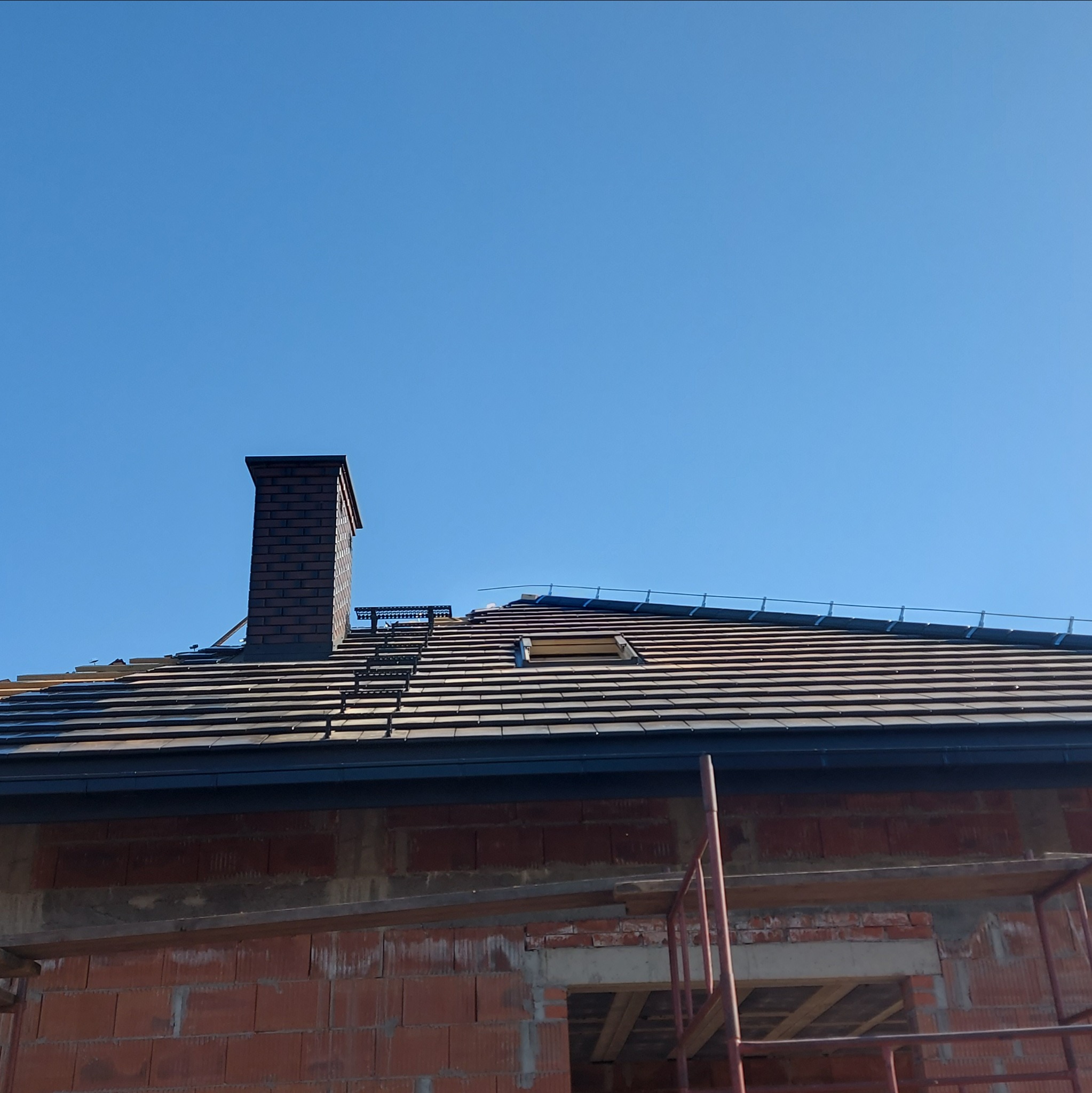 Ceglany komin na dachu z brązową dachówką, widoczna drabinka dachowa i fragment muru w surowym stanie. Błękitne niebo w tle.