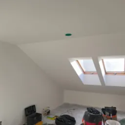 Pok&oacute;j na poddaszu po szpachlowaniu i malowaniu, widoczne okna dachowe, narzędzia i odkurzacz przemysłowy. Świeżo pomalowane ściany i sufit na biało.
