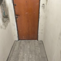 Paweł Sawicki - Korytarz w trakcie remontu: brązowe drzwi, jasne ściany przed malowaniem, szare płytki imitujące drewno na podłodze. Widoczny interkom i niedokończone obramowanie otworu.