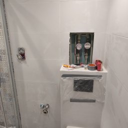 Paweł Sawicki - Białe płytki w łazience w Płocku, z widocznymi licznikami wody i narzędziami. Ślady po pracach wykończeniowych, widoczna kabina prysznicowa i instalacja.