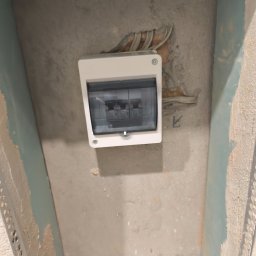 Paweł Sawicki - Biała skrzynka elektryczna z bezpiecznikami zamontowana na szarej, surowej ścianie w trakcie remontu. Widoczne kable i ślady szpachlowania.