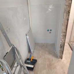 Biała łazienka w trakcie remontu: świeżo położone płytki z falistym wzorem, drabina malarska, narzędzia malarskie i płyta gipsowa.