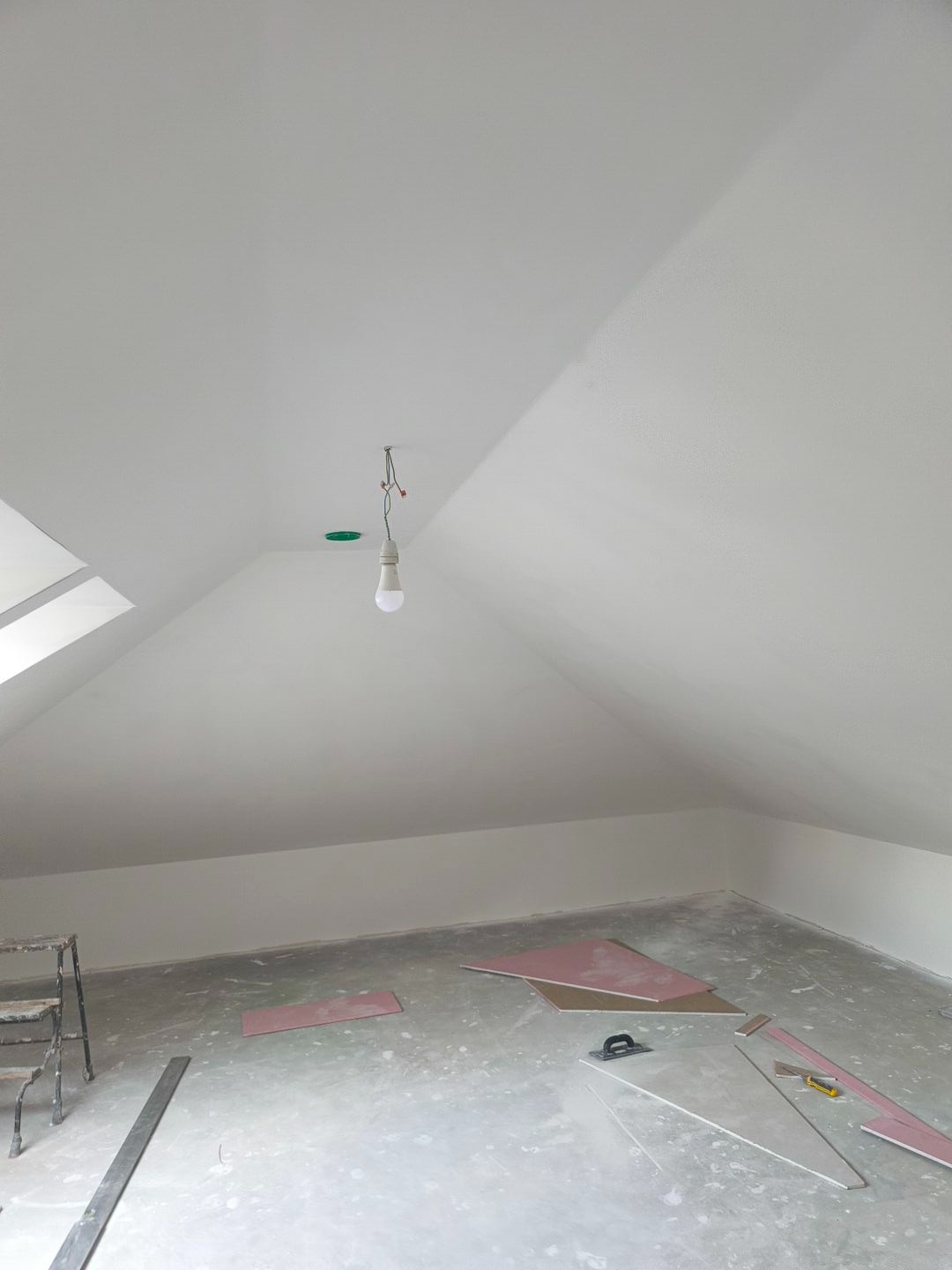 Poddasze w trakcie szpachlowania: białe ściany, płyty gipsowe, narzędzia glazurnicze i drabina. Oświetlenie tymczasowe, okno dachowe po lewej.
