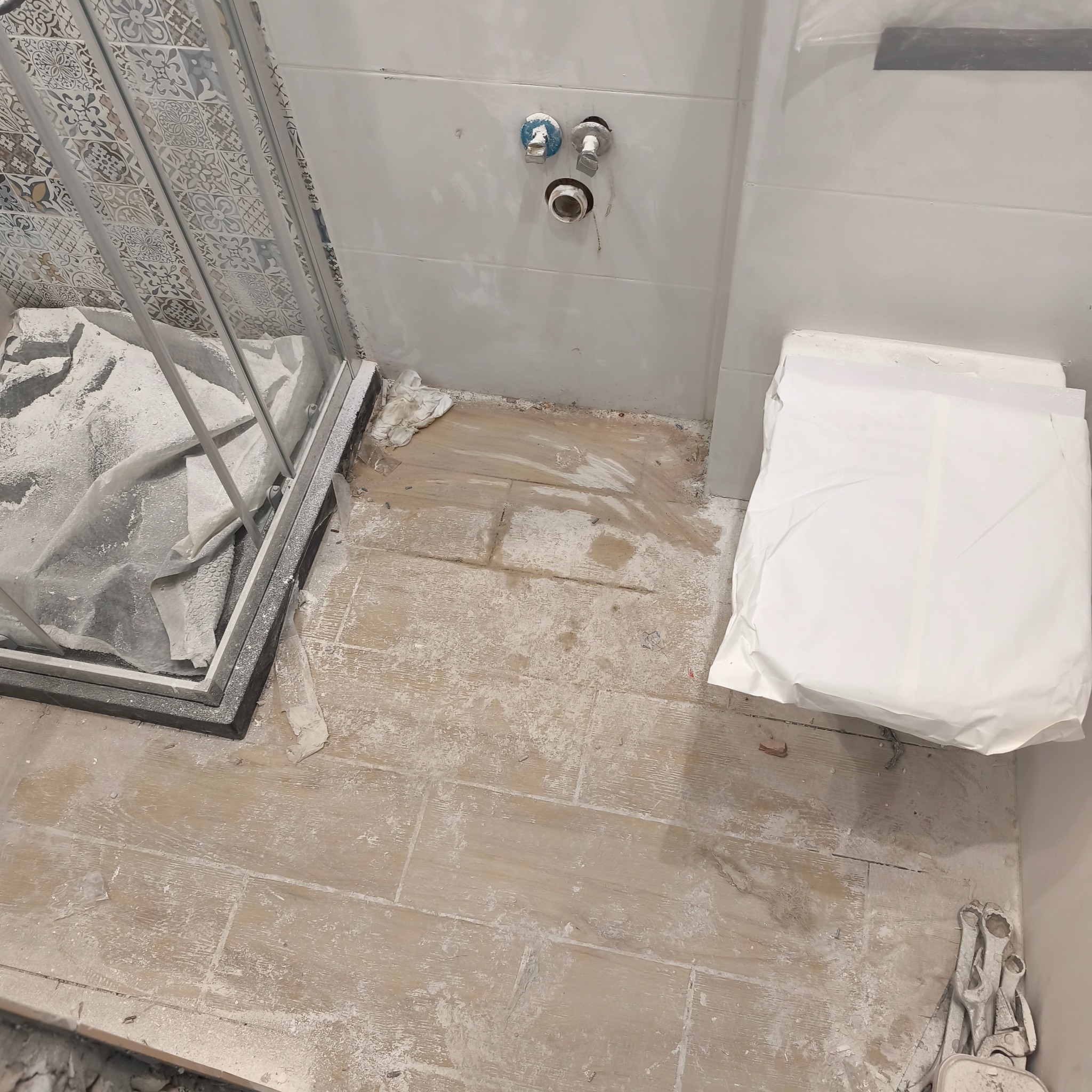 Remontowana łazienka z zabezpieczoną kabiną prysznicową i toaletą. Widoczne płytki imitujące drewno na podłodze i przygotowane przyłącza wodne na ścianie.