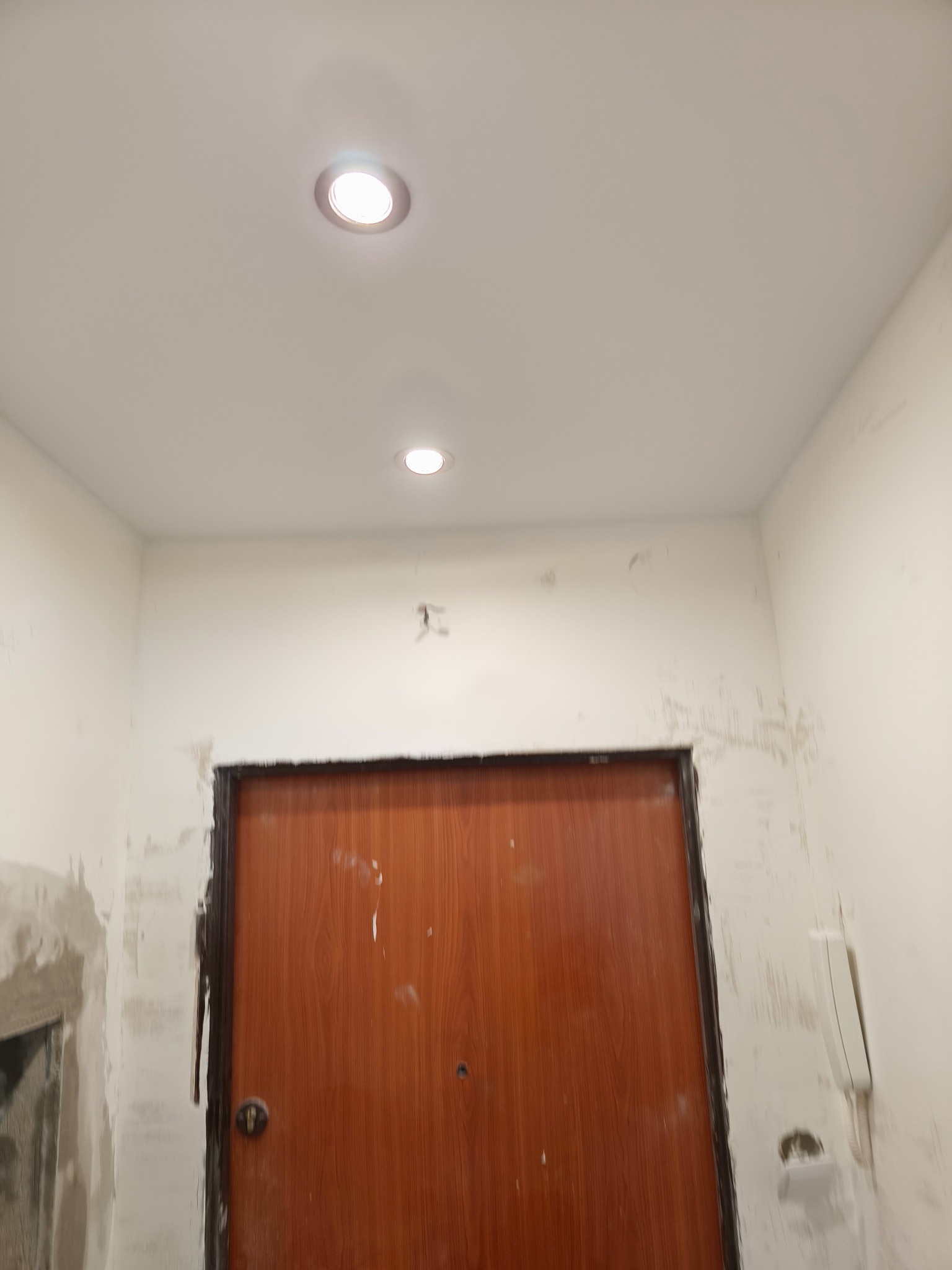 Wnętrze w trakcie prac: ściany po szpachlowaniu, widoczne ślady po gładzi, zamontowane oświetlenie sufitowe i drzwi. Przygotowanie do dalszych prac wykończeniowych.