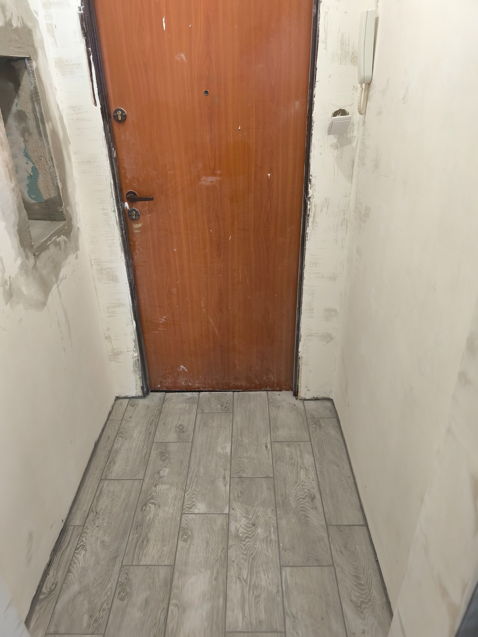 Korytarz w trakcie remontu: brązowe drzwi, jasne ściany przed malowaniem, szare płytki imitujące drewno na podłodze. Widoczny interkom i niedokończone obramowanie otworu.