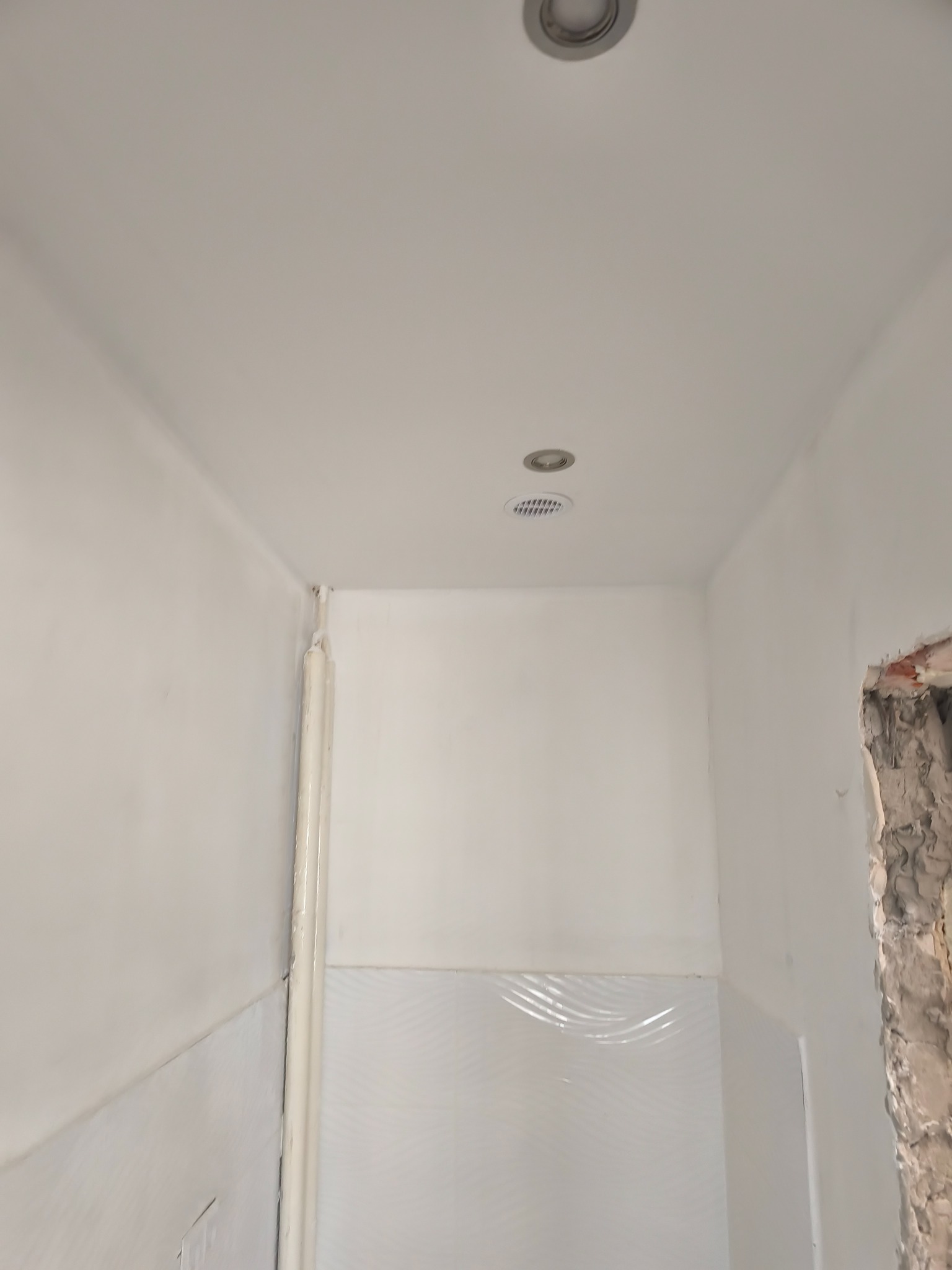 Wnętrze łazienki w trakcie prac wykończeniowych: białe ściany z częściowo położonymi płytkami, widoczne rury i sufit z dwoma punktami oświetleniowymi.