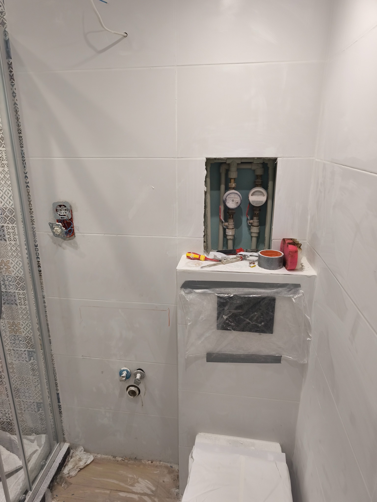 Białe płytki w łazience w Płocku, z widocznymi licznikami wody i narzędziami. Ślady po pracach wykończeniowych, widoczna kabina prysznicowa i instalacja.
