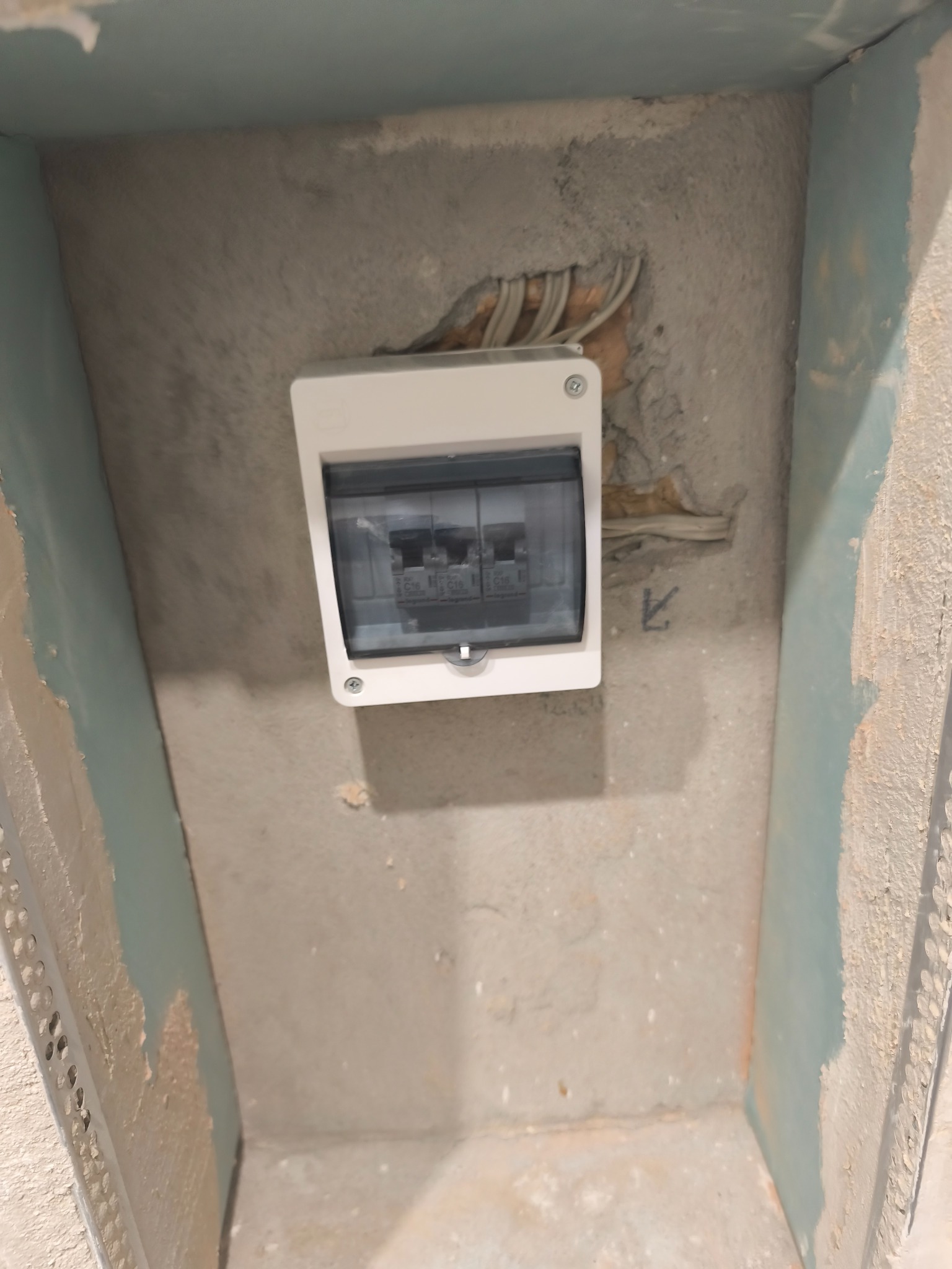 Biała skrzynka elektryczna z bezpiecznikami zamontowana na szarej, surowej ścianie w trakcie remontu. Widoczne kable i ślady szpachlowania.