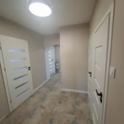 Jasne wnętrze korytarza z beżowymi ścianami, wzorzystą podłogą i białymi drzwiami z czarnymi klamkami. Widok na oświetlony sufit i dalszą część korytarza.