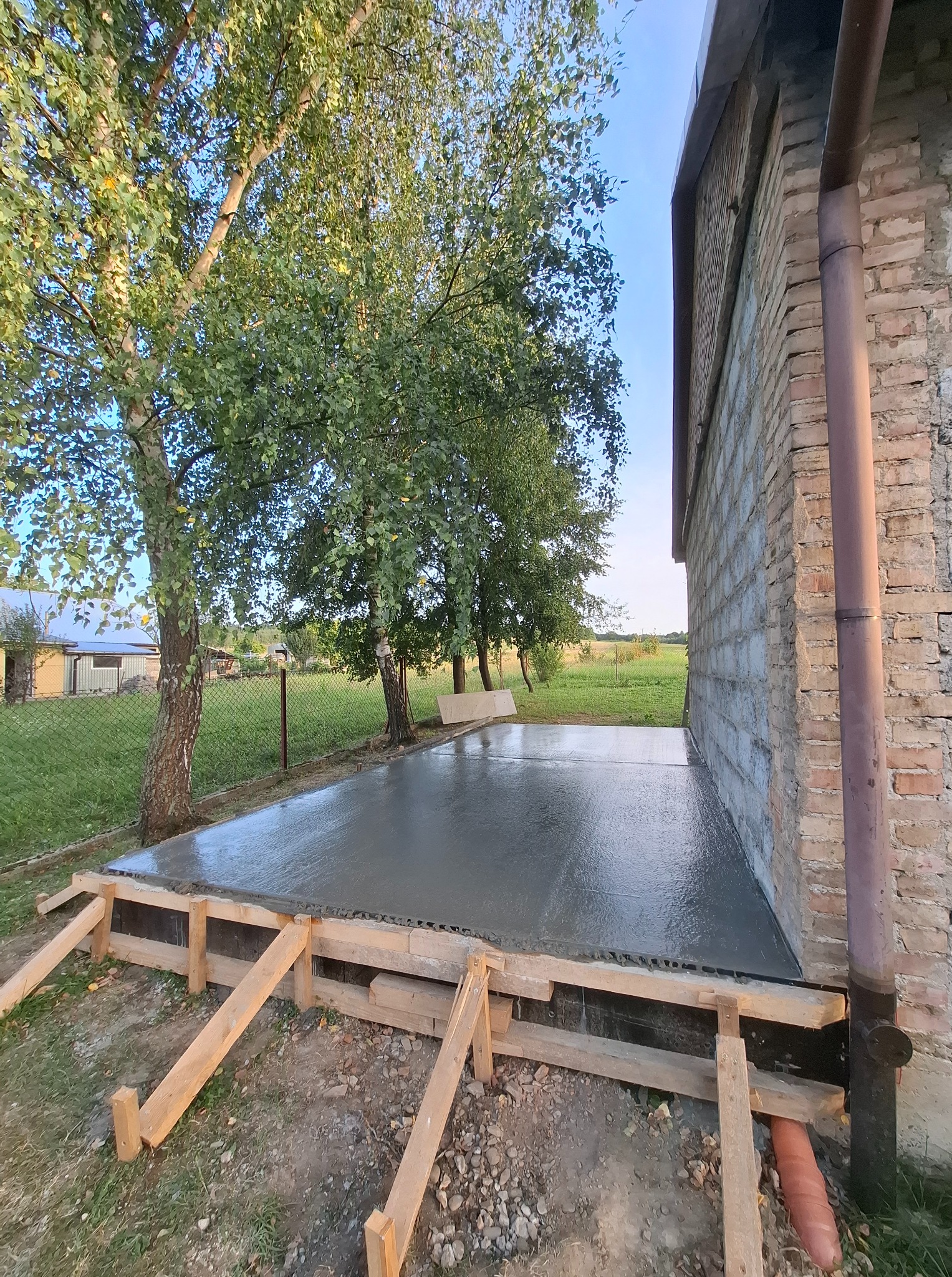 Świeżo wylana, gładka wylewka betonowa w drewnianym szalunku, przylegająca do ściany budynku z cegły, na tle zieleni i błękitnego nieba. Widoczne drzewa i trawa.