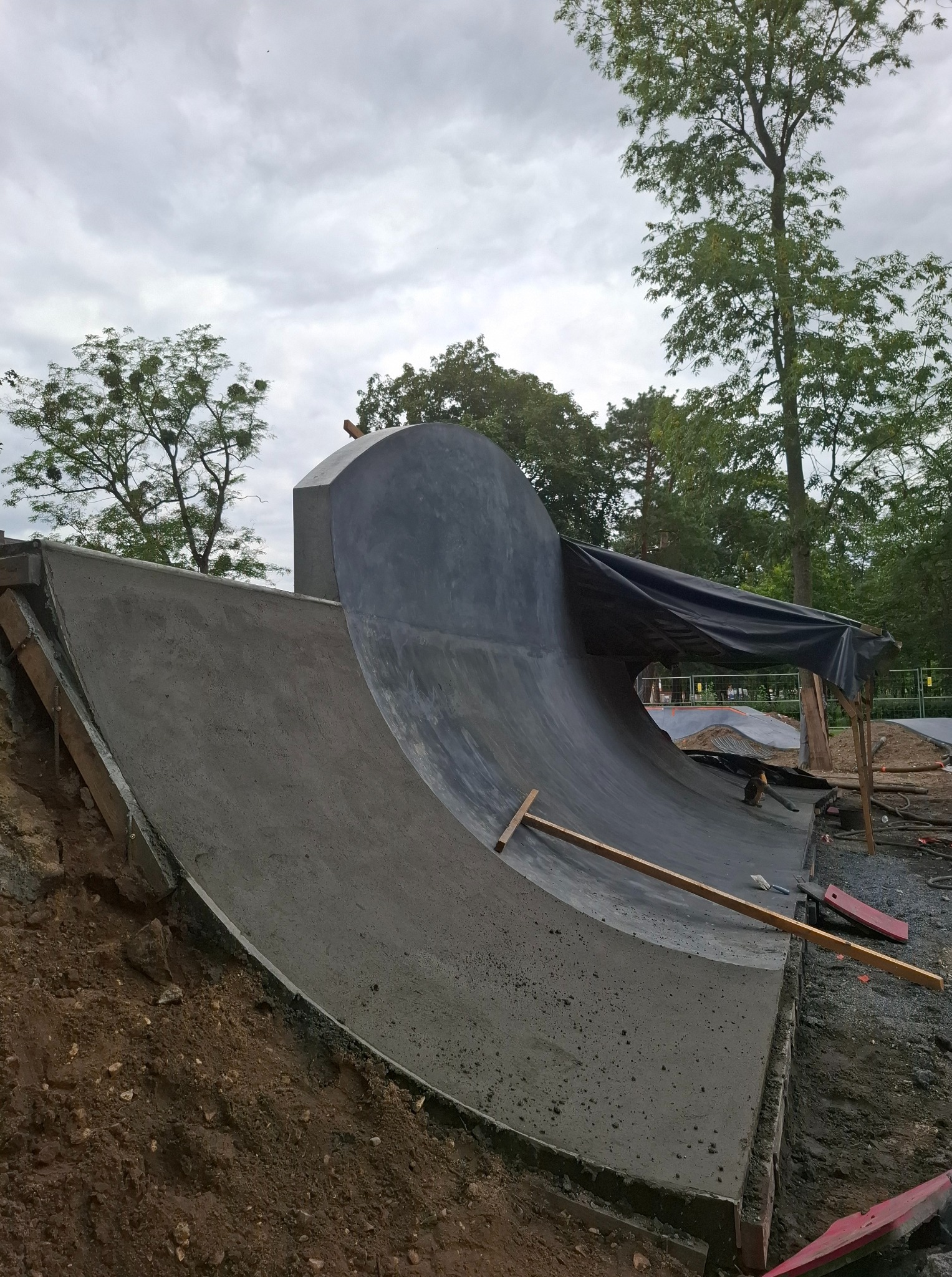 Budowa betonowego skateparku w Tarnowcu: świeżo wylany element z drewnianym stemplem, widoczne zbrojenie i teren budowy w tle. Prace w toku.