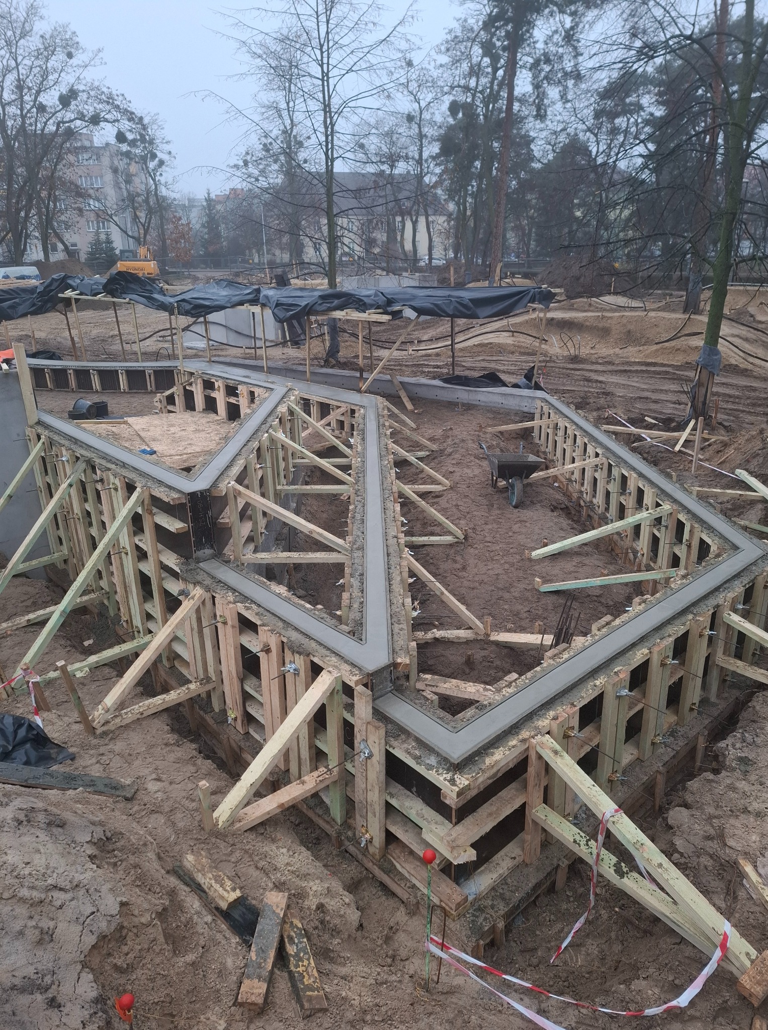 Zalewanie fundamentów pod budowę, widoczne drewniane szalunki wypełnione świeżym betonem. W tle drzewa i budynki w pochmurny dzień.