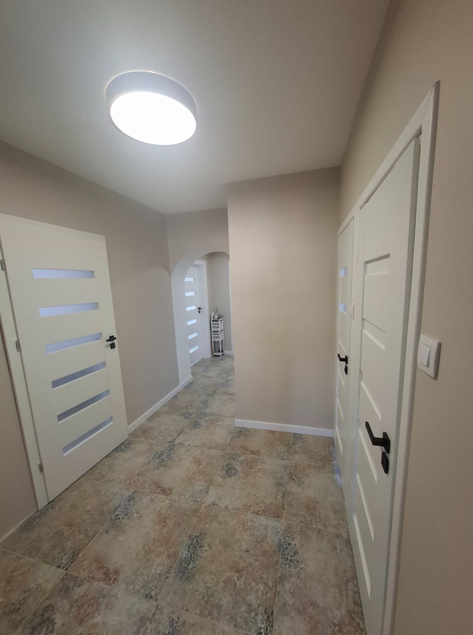 Jasne wnętrze korytarza z beżowymi ścianami, wzorzystą podłogą i białymi drzwiami z czarnymi klamkami. Widok na oświetlony sufit i dalszą część korytarza.