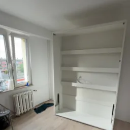 Biała, pusta szafa wnękowa z p&oacute;łkami, zamontowana w jasnym pokoju z oknem i grzejnikiem. Widoczne mechanizmy otwierania. Podłoga wyłożona panelami.