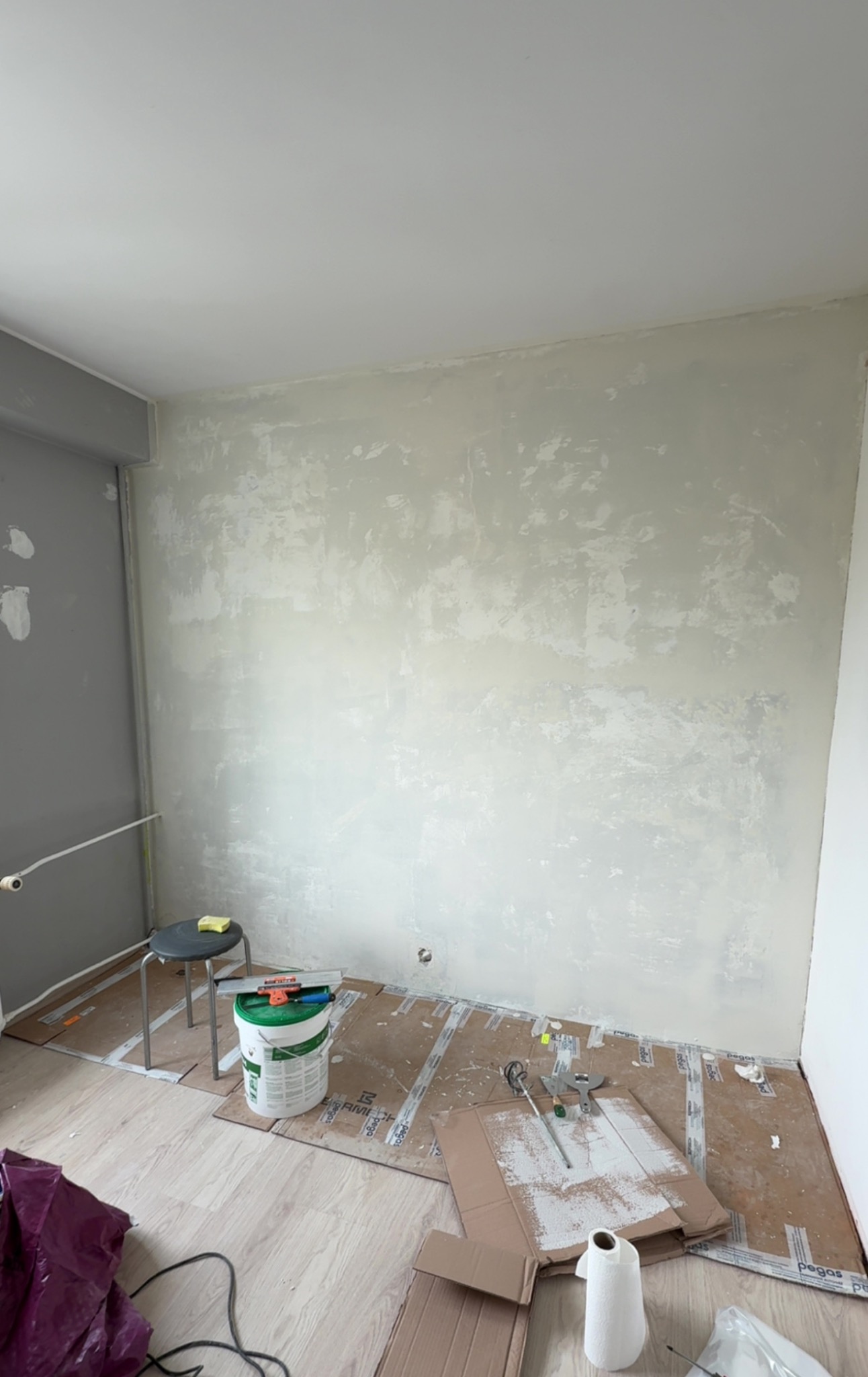 Ściana po szpachlowaniu, przygotowana do malowania. Widoczne narzędzia i materiały budowlane. Podłoga zabezpieczona kartonem. Krzesło z gąbką stoi obok ściany.