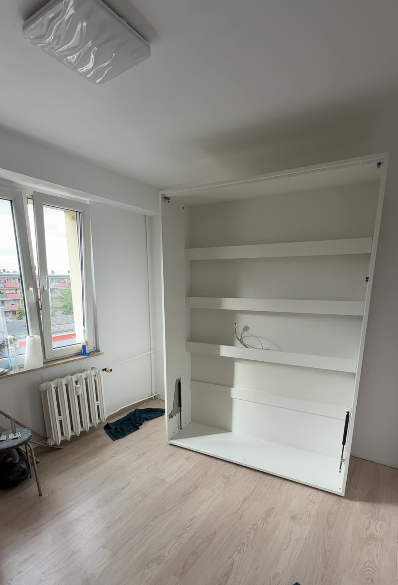 Biała, pusta szafa wnękowa z półkami, zamontowana w jasnym pokoju z oknem i grzejnikiem. Widoczne mechanizmy otwierania. Podłoga wyłożona panelami.