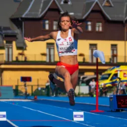 Kobieta w locie podczas skoku w dal na zawodach lekkoatletycznych. Widoczny numer startowy i sponsorzy. W tle budynek i ambulans.