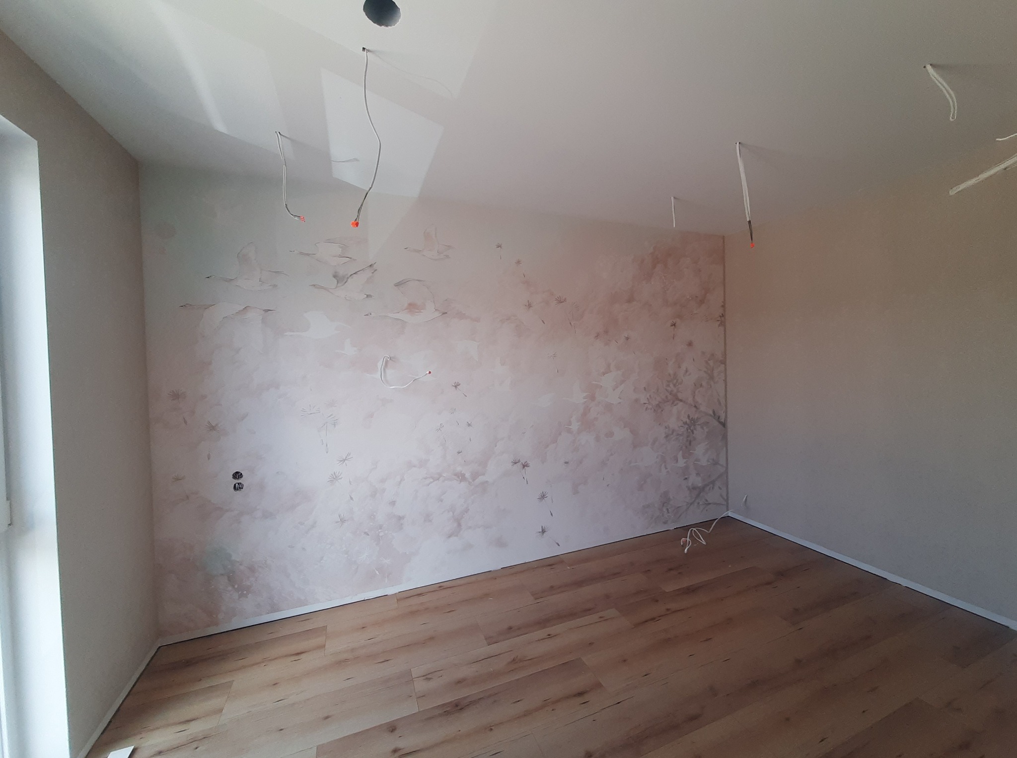 Pokój po remoncie: parkiet w odcieniach brązu, ściana z motywem ptaków i drzew w tonacji beżowo-brązowej. Widoczne wystające kable z sufitu.