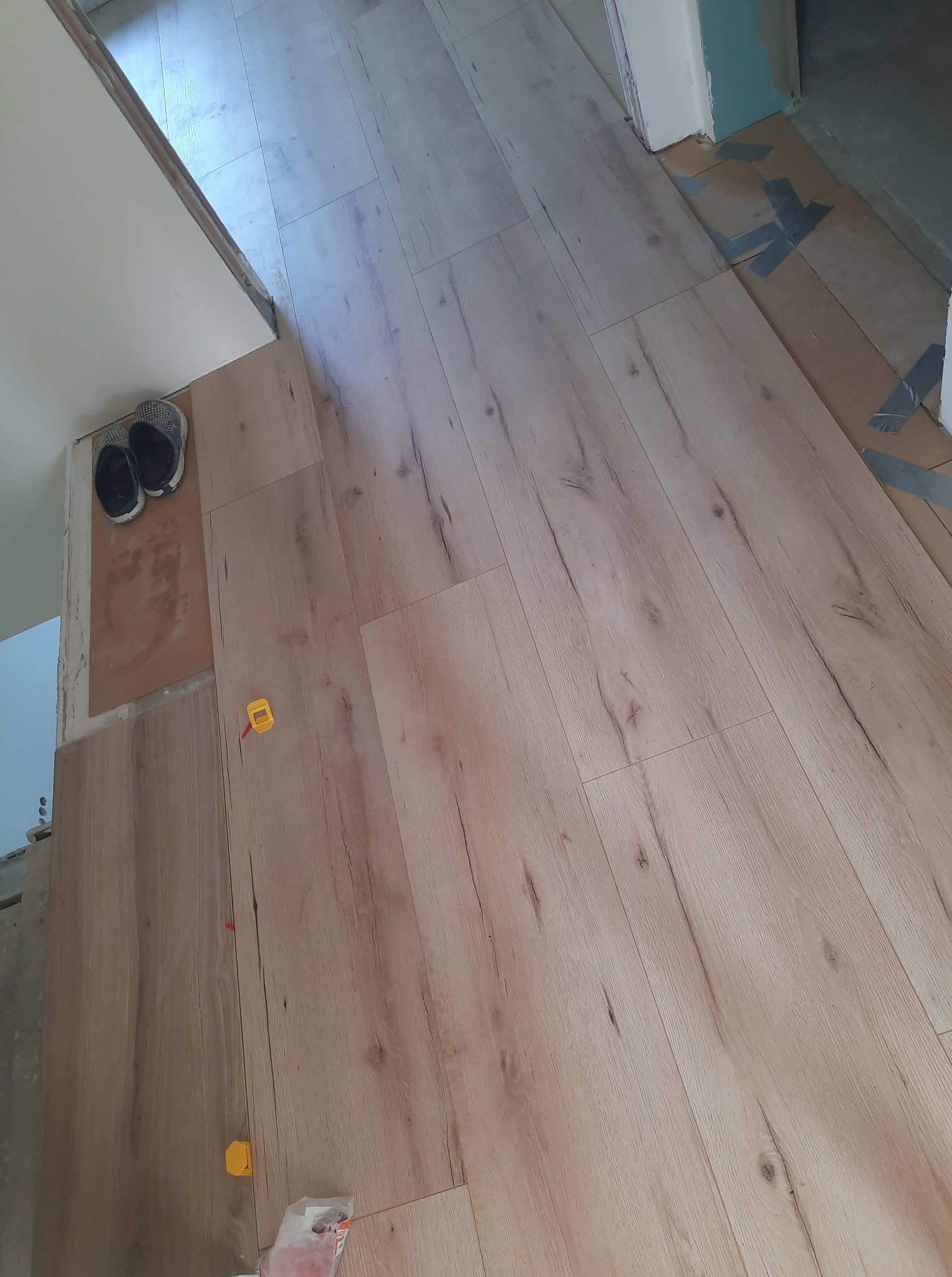 Układanie paneli podłogowych z widocznymi narzędziami, w tle buty robocze i przygotowane podłoże, etap prac wykończeniowych.