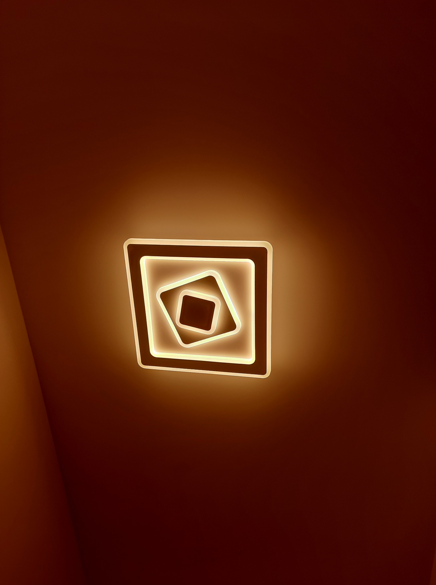 Nowoczesna lampa sufitowa LED w kształcie kwadratów, emitująca ciepłe, żółte światło na tle ciemnego sufitu. Geometryczny design oświetlenia.