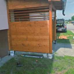 Drewniana altana przy budynku, częściowo zabudowana, z widocznymi narzędziami i elementami konstrukcyjnymi w trakcie budowy. W tle zaparkowany samochód.