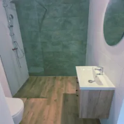 Nowoczesna łazienka po remoncie: kabina prysznicowa z zielonymi płytkami, drewnopodobne panele podłogowe i szafka z umywalką, okrągłe lustro.