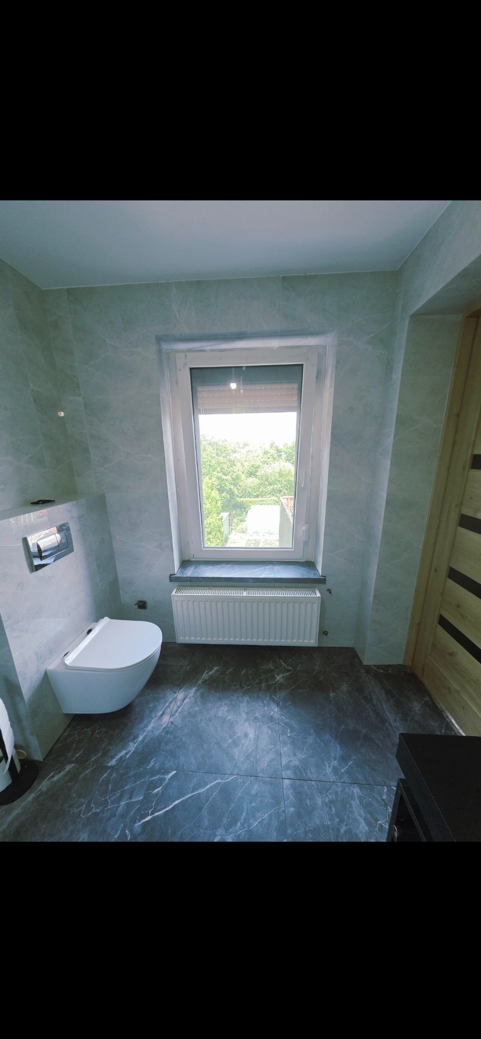 Nowoczesna łazienka z szarymi marmurowymi płytkami na ścianach i podłodze. Podwieszana toaleta i okno z widokiem na zieleń.