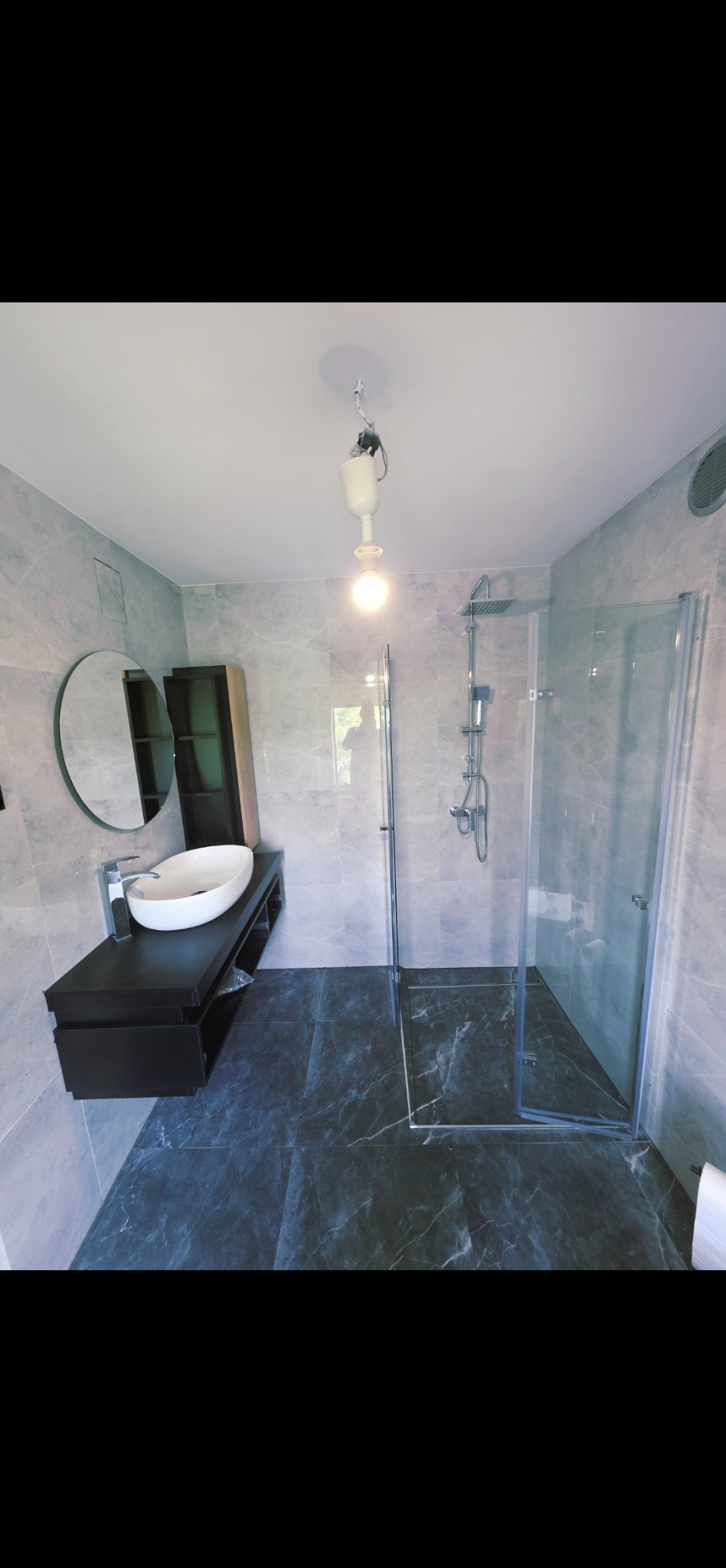Stylowa łazienka z szarymi marmurowymi płytkami, kabiną prysznicową i umywalką na czarnej szafce. Widoczne okrągłe lustro i niedokończona lampa sufitowa.