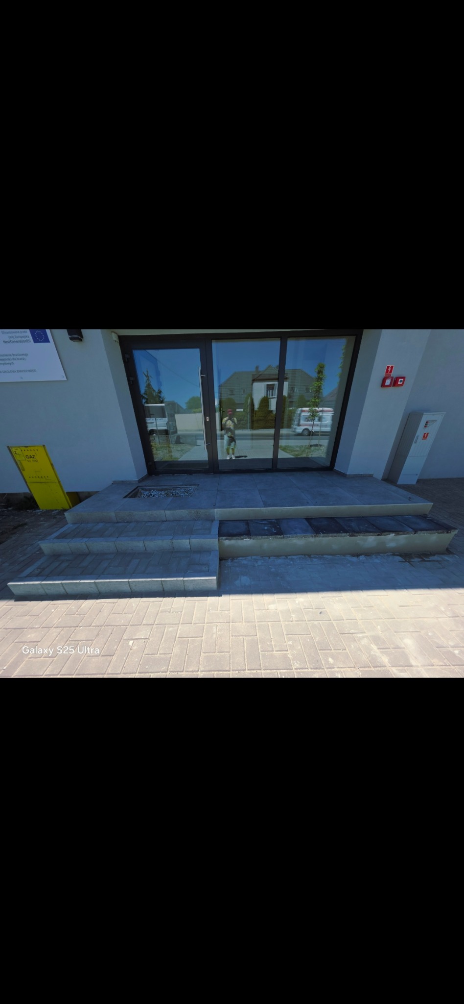 Układanie kostki brukowej na schodach wejściowych do budynku. Widoczne świeżo położone stopnie i chodnik z kostki. W tle drzwi wejściowe z odbiciem otoczenia.