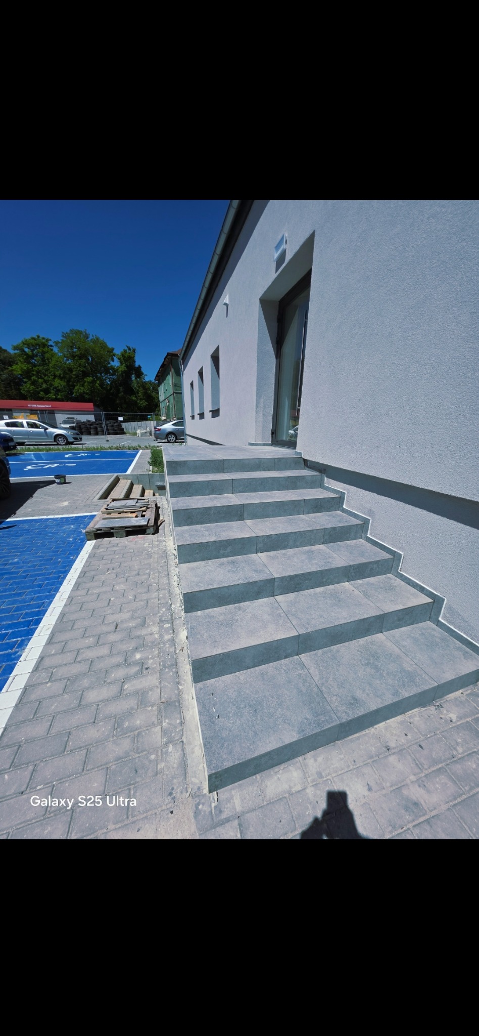 Nowe, szare płytki schodowe prowadzą do budynku z białymi ścianami. Widoczne zaparkowane samochody i fragment niebieskiego parkingu dla niepełnosprawnych.