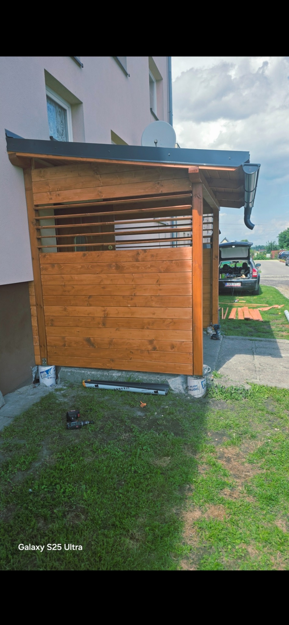 Drewniana altana przy budynku, częściowo zabudowana, z widocznymi narzędziami i elementami konstrukcyjnymi w trakcie budowy. W tle zaparkowany samochód.
