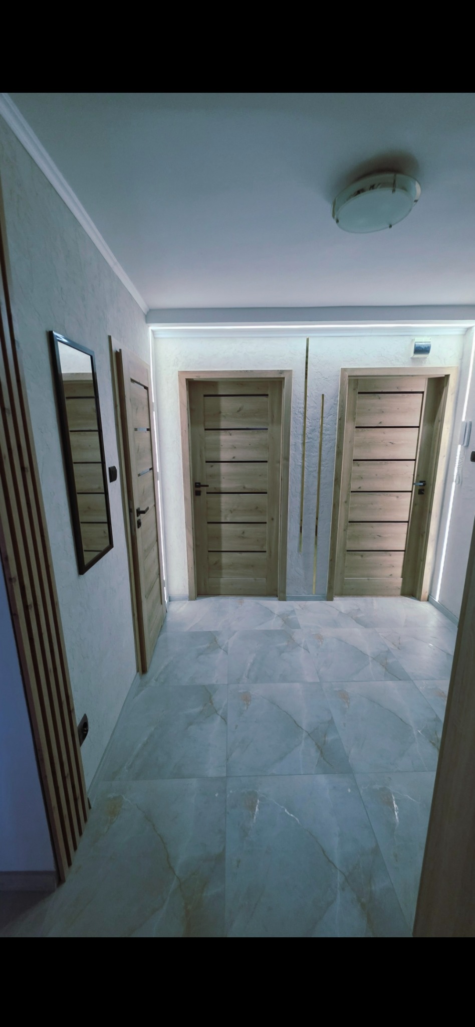 Wykończony korytarz z dwiema drewnianymi drzwiami, lustrem i dekoracyjnymi lamelami na ścianie. Podłoga wyłożona płytkami imitującymi marmur.