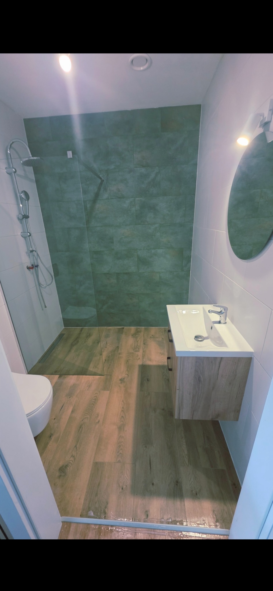 Nowoczesna łazienka po remoncie: kabina prysznicowa z zielonymi płytkami, drewnopodobne panele podłogowe i szafka z umywalką, okrągłe lustro.