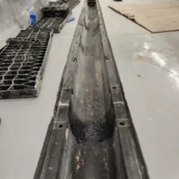 Ukończony montaż odpływu liniowego w posadzce, widoczne elementy rusztu. Szara posadzka, czarne elementy odpływu. Ujęcie z góry, perspektywa zbieżna.