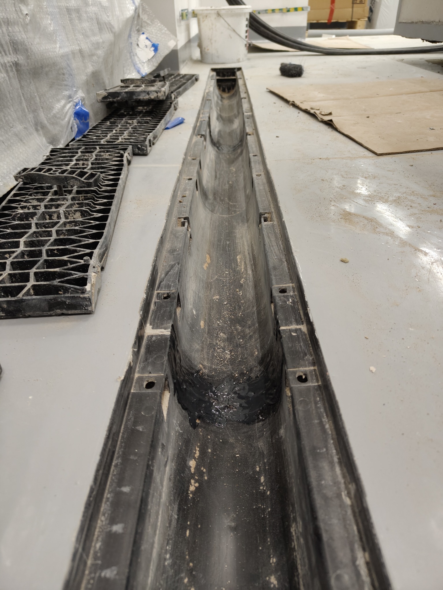 Ukończony montaż odpływu liniowego w posadzce, widoczne elementy rusztu. Szara posadzka, czarne elementy odpływu. Ujęcie z góry, perspektywa zbieżna.