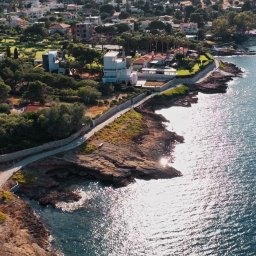 Adam Żurawicki - Panoramiczny widok z lotu ptaka na nadmorską miejscowość z domami, zielenią i błękitnym morzem w słoneczny dzień. Widoczna linia brzegowa i ścieżka wzdłuż klifu.