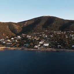 Zdjęcie z lotu ptaka przedstawia nadmorskie miasteczko z białymi domami na zboczu góry, z anteną na szczycie. W dole ciemna woda i błękitne niebo.