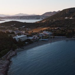 Adam Żurawicki - Panoramiczny widok z lotu ptaka na nadmorski kurort z plażą, hotelem i gęstą roślinnością w tle, uchwycony w miękkim świetle wczesnego ranka lub późnego popołudnia.