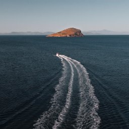 Adam Żurawicki - Błękitne morze z motorówką zostawiającą ślad piany, w tle górzyste wyspy i żaglówki, słoneczny dzień.