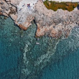 Adam Żurawicki - Widok z góry na krystalicznie czystą wodę obmywającą skalisty brzeg z osobami odpoczywającymi na ręczniku i pływającą osobą, uchwycony w słoneczny dzień.