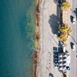 Adam Żurawicki - Widok z lotu ptaka na turkusowe morze, piaszczystą plażę z parasolami, drogę z samochodami i budynki. Ujęcie z góry ukazuje detale wybrzeża i zabudowy.