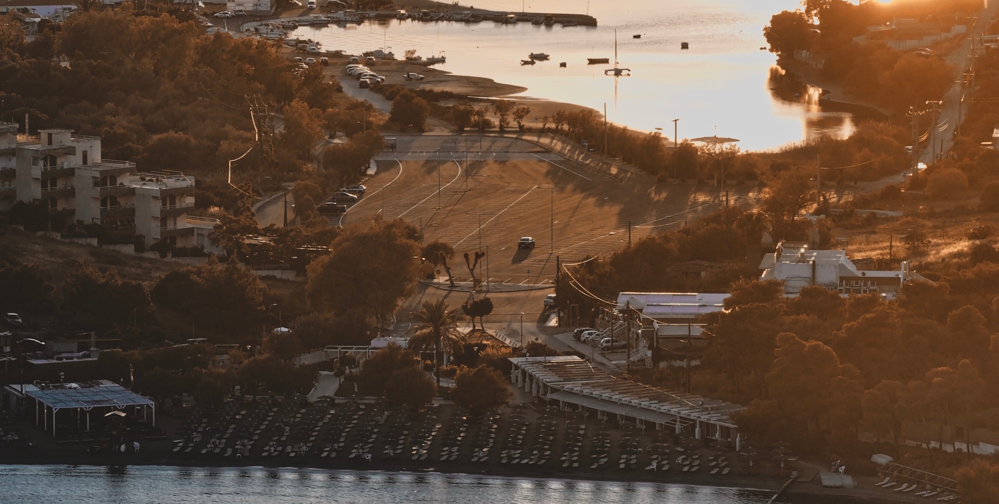 Widok z góry na plażę z leżakami, parking z jednym samochodem i budynki otoczone drzewami w ciepłym, zachodzącym słońcu nad morzem.
