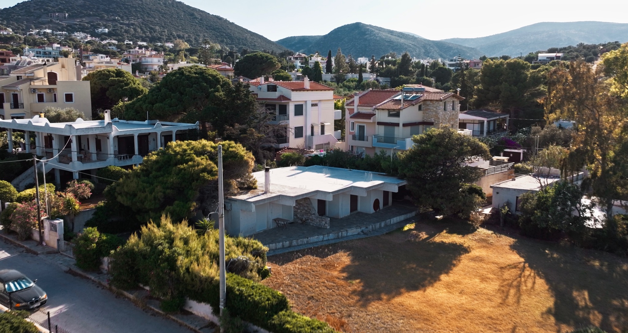 Zdjęcie z drona przedstawia osiedle domów jednorodzinnych z widokiem na góry. W kadrze dom z białym dachem i kamiennym wykończeniem, otoczony zielenią i suchą trawą.