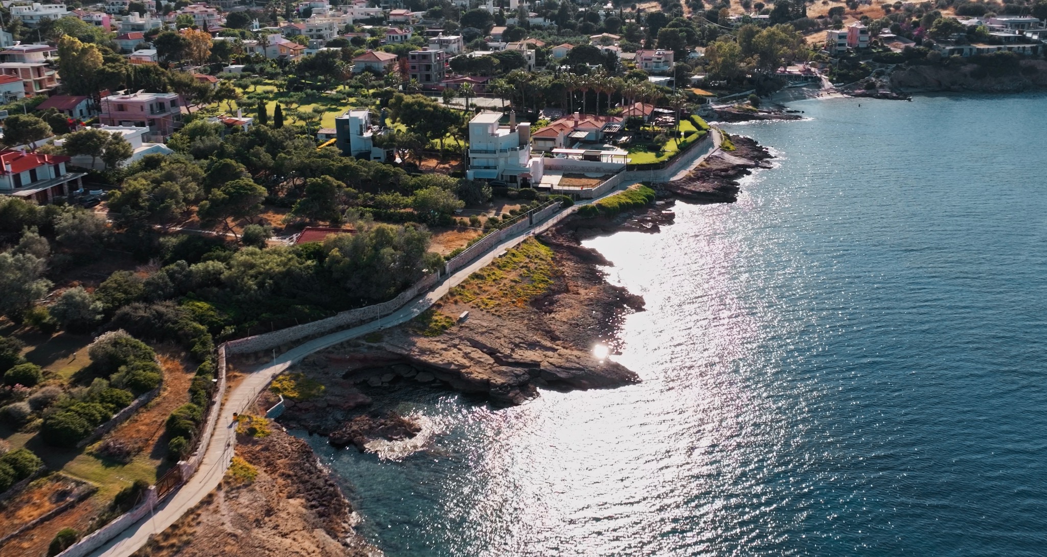 Panoramiczny widok z lotu ptaka na nadmorską miejscowość z domami, zielenią i błękitnym morzem w słoneczny dzień. Widoczna linia brzegowa i ścieżka wzdłuż klifu.